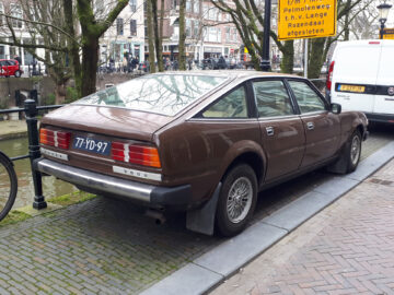 Een bruine, vintage Rover 3500 vierdeursauto uit 1977 staat geparkeerd in een bakstenen straat naast een gracht. Op het kenteken van de auto staat "77-YD-97." Op de achtergrond zijn er bomen en gebouwen.