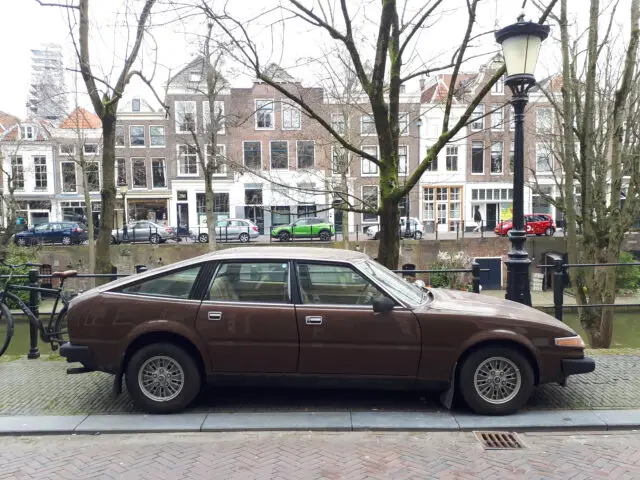 Un Rover 3500 vintage marrón está aparcado en un canal, delante de una hilera de edificios de ladrillo y árboles. El cielo está nublado y se ve una farola a la derecha.