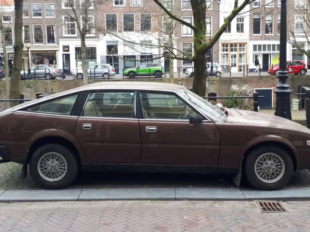 Een bruine Rover 3500 uit 1977 staat geparkeerd in een geplaveide straat naast een gracht, met fietsen en oude gebouwen zichtbaar op de achtergrond. #Gespot