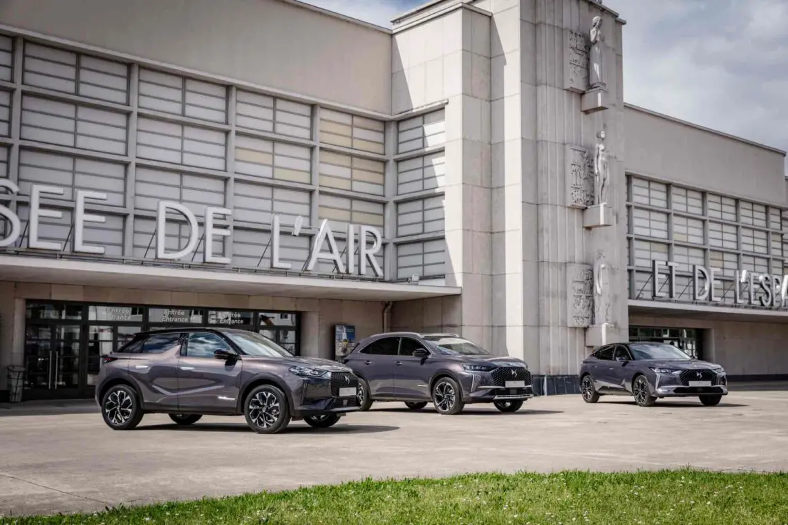 Drie donkergekleurde SUV's staan geparkeerd voor een gebouw met grote ramen en de woorden "MUSEE DE L'AIR" op de gevel. De lucht is gedeeltelijk bewolkt en vormt een dramatisch decor voor de onthulling door DS onthult van een exclusieve collectie ter ere van Antoine de Saint-Exupéry.
