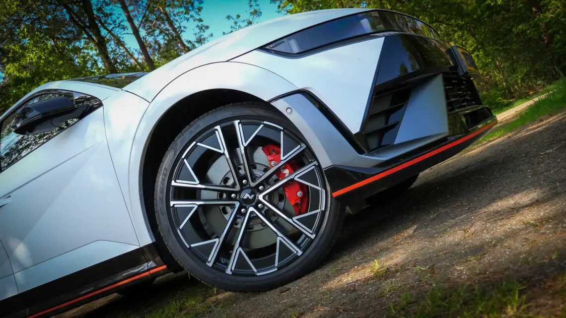 Shot vanuit een lage hoek van de voorkant van een moderne witte Hyundai Ioniq met opvallende zwart-zilveren lichtmetalen velgen, rode remklauwen en strakke carrosserielijnen, tegen een bosrijke achtergrond.