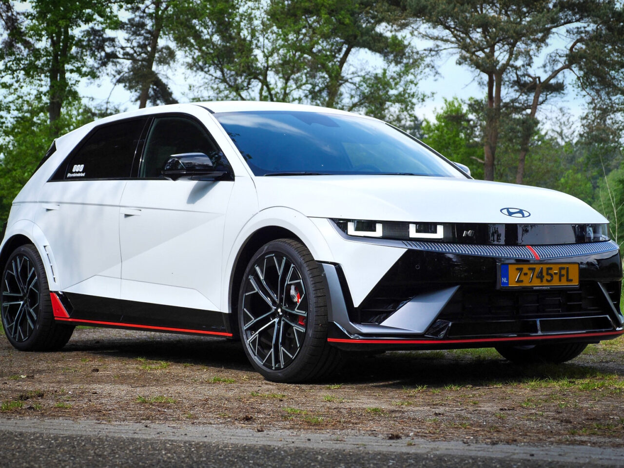 Een witte elektrische auto Hyundai Ioniq 5 N met zwarte accenten en rode details, geparkeerd op een landelijke weg omgeven door bomen en groen. Het strakke design van de Hyundai Ioniq valt op tegen de serene achtergrond van de natuur.