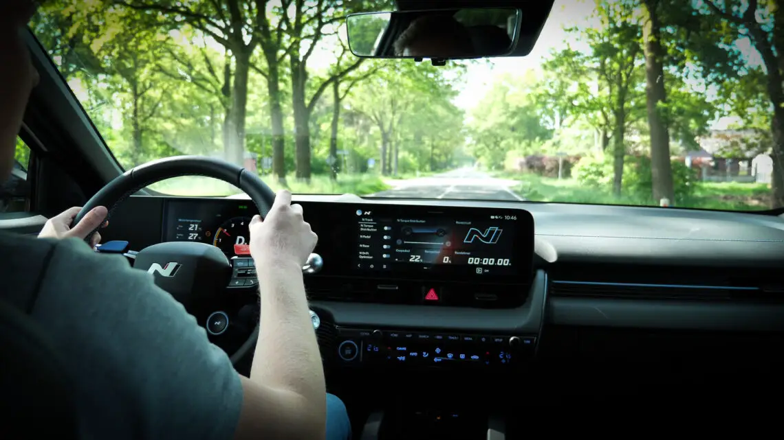 Een persoon rijdt in een Hyundai Ioniq over een met bomen omzoomde weg. Het dashboard en het stuur van de auto zijn zichtbaar en geven navigatie- en voertuiginformatie weer.
