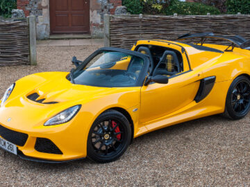 Coche deportivo Lotus Exige S amarillo brillante aparcado delante de un edificio rústico con vallas tejidas.