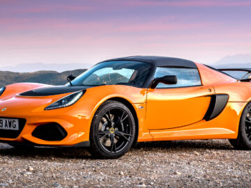 Oranje Lotus Exige S sportwagen geparkeerd op een heuvel bij zonsondergang met schilderachtige bergachtergrond.