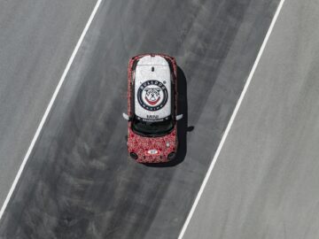 Een MINI John Cooper Works-auto met een wit en rood patroon en een Bulldog Racing-logo rijdt op een rijbaan. De auto is van bovenaf bekeken.