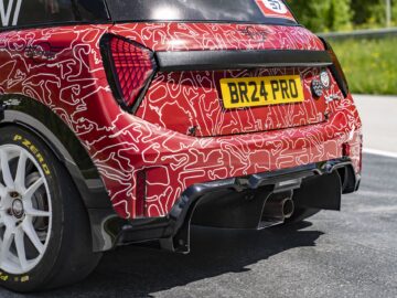 Een close-up van de achterkant van een rode raceauto, die lijkt op een MINI John Cooper Works, met abstracte ontwerpen en een gele kentekenplaat met de tekst 'BR24 PRO'. Hij is voorzien van speciale Pirelli-banden en staat geparkeerd op een verharde weg met groene vegetatie op de achtergrond, wat de triomfantelijke terugkeer symboliseert.