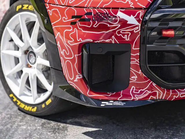 Close-up van de rechtervoorkant van een raceauto met een rood-witte verflaag, banden met het merk Pirelli en het merk MINI John Cooper Works. De auto lijkt op een verhard oppervlak geparkeerd te staan.