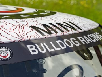 Close-up van de voorruit van een raceauto met het merk 'Bulldog Racing' en een logo met een bulldog-gezicht. Op het dak zijn de letters "MINI John Cooper Works" zichtbaar.