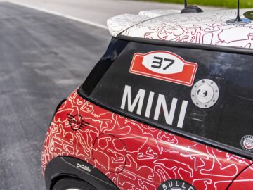 Close-up van de achterruit van een rood-witte auto, met het nummer 37 en het woord "MINI John Cooper Works" in vetgedrukte letters. Het model uit 2023 heeft een uniek rood-wit camouflagepatroon en een zilveren tankdop, wat zijn indrukwekkende terugkeer markeert.