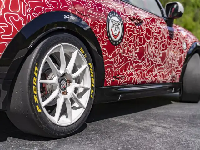 Zijaanzicht van een rode MINI John Cooper Works met witte abstracte ontwerpen, met een bulldog-logo en uitgerust met Pirelli P Zero-banden op witte wielen, geparkeerd op een verhard terrein met groen op de achtergrond.