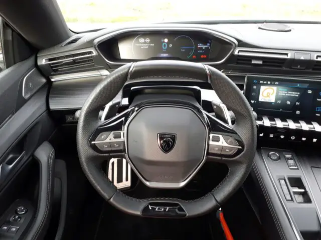 Binnenaanzicht van de Peugeot 508 uit 2024 met het stuur, het digitale dashboard en het touchscreen-display op de middenconsole. De voertuigbedieningen zijn verbeterd met knoppen en knoppen voor gebruikersgemak.