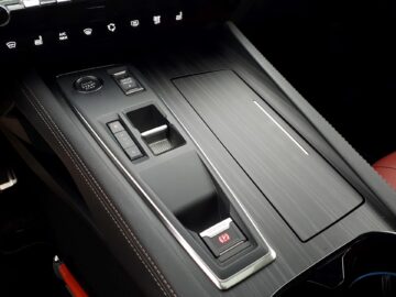 Close-up van de middenconsole van een Peugeot 508 SW Hybrid met verschillende bedieningselementen, waaronder een versnellingspook, een elektronische parkeerremknop en meerdere andere knoppen. Het strakke, moderne design wordt benadrukt in deze review uit 2024.