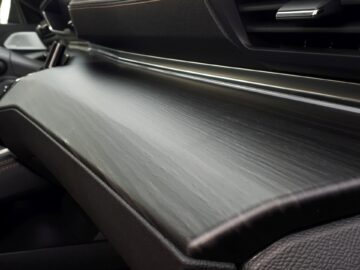Een close-up van het dashboard van de Peugeot 508 SW uit 2024 met een strakke, matzwarte textuur met subtiele nerfpatronen, geaccentueerd door donker, gestikt leer en een chromen strip. Het Hybrid 225-model toont een innovatief ontwerp en milieuvriendelijke technologie.
