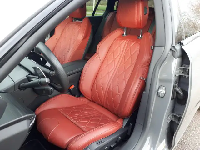 Een Peugeot 508 SW-interieur met twee voorstoelen met roodleren bekleding en diamantstiksel, een zwart stuur en een open bestuurdersdeur. Dit Hybride 225-model uit 2024 combineert luxe met de allernieuwste technologie.