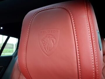 Close-up van de hoofdsteun van een roodleren autostoel met het in reliëf gemaakte Peugeot-logo met een leeuw in een schild, onderdeel van het strakke ontwerp van de nieuwe Peugeot 508 SW Hybrid.