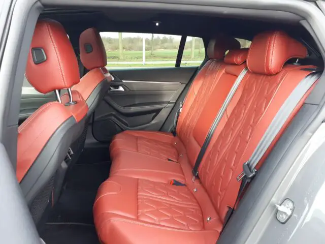 Binnenaanzicht van de Peugeot 508 SW uit 2024 met de achterstoelen, die zijn bekleed met rood leer met gedetailleerde stiksels. De autodeur staat open en de stoelen zien er schoon en goed onderhouden uit, wat het verfijnde ontwerp van dit Hybrid 225-model benadrukt.
