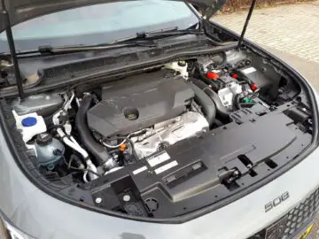 Er is een automotor zichtbaar met de motorkap open, waardoor verschillende componenten zichtbaar zijn in de motorruimte van een Peugeot 508 SW Hybrid 225 uit 2024, geparkeerd op een verhard oppervlak.
