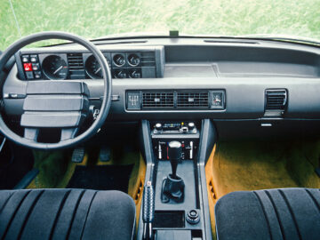 Innenraum eines Rover 3500 von 1977 mit Armaturenbrett, Lenkrad, Schalthebel und Mittelkonsole, mit Blick durch die Windschutzscheibe vom Gras aus. Gesichtet.