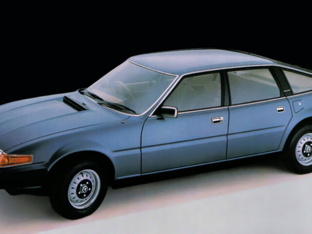 Een blauwe vintage sedan Rover 3500 uit 1977 met hatchback-ontwerp staat geparkeerd in een studioomgeving tegen een zwarte achtergrond. De auto beschikt over vier deuren en lichtmetalen velgen.