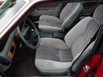 L'intérieur de la Rover 3500 de 1977 montre deux sièges avant avec des garnitures grises, le volant, le tableau de bord et la console centrale, avec la porte du conducteur ouverte.