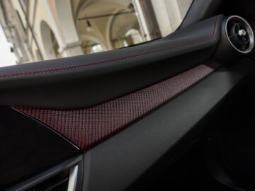 Close-up van het dashboard van een auto met zwarte en rode stiksels, een gestructureerde afwerking en een emblematisch Quadrifoglio-embleem. Een ventilatierooster en een deel van het infotainmentscherm zijn zichtbaar en stralen de sportieve elegantie uit die synoniem is aan Alfa Romeo.