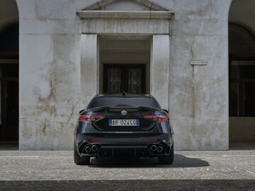 Een zwarte Alfa Romeo Giulia Quadrifoglio staat geparkeerd voor een witstenen gebouw met structuur en gebogen deuropeningen en ramen. Op het kenteken staat "AR 024GQ".