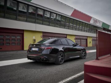 Een zwarte Alfa Romeo sedan staat geparkeerd op een racecircuit naast rode en witte garagegebouwen onder een bewolkte hemel. De auto, mogelijk een Quadrifoglio- of Super Sport-model, beschikt over dubbele uitlaten en is voorzien van een kentekenplaat.