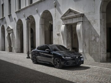 Een strakke zwarte Alfa Romeo Quadrifoglio sedan staat geparkeerd in een geplaveide straat naast een gebroken wit gebouw met boogramen en deuropeningen.