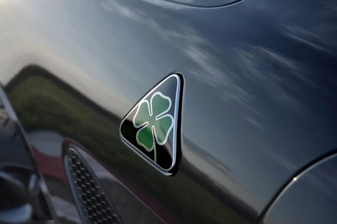 Close-up van het metallic zwarte oppervlak van een auto met een driehoekig embleem met een groen klavertje vier, emblematisch voor Alfa Romeo's Quadrifoglio Super Sport-erfenis.