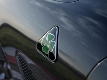 Close-up van het metallic zwarte oppervlak van een auto met een driehoekig embleem met een groen klavertje vier, emblematisch voor Alfa Romeo's Quadrifoglio Super Sport-erfenis.