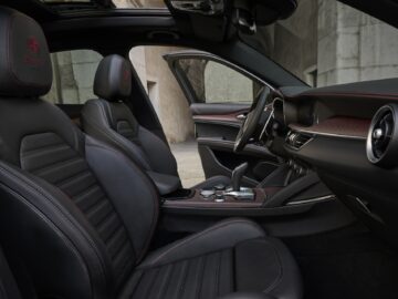 Interieur van een moderne Alfa Romeo met zwartleren stoelen met rode stiksels, een middenconsole met bedieningselementen en een gedeeltelijk open schuifdak, tegen een achtergrond van betonnen muren. De fijne details van de Quadrifoglio stralen luxe en sportiviteit uit.