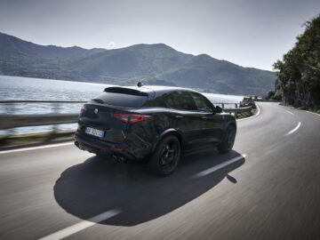 We zien een zwarte SUV, mogelijk een Alfa Romeo Quadrifoglio, rijden op een bochtige weg naast een grote watermassa met bergen op de achtergrond.