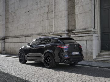 Een zwarte luxe SUV, mogelijk een Alfa Romeo Quadrifoglio, staat geparkeerd in een geplaveide straat naast een groot stenen gebouw. Op het kenteken van het voertuig staat "AR 002 JQ".