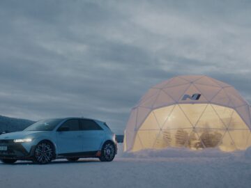 Een blauwe Hyundai IONIQ 5 N staat in de sneeuw geparkeerd naast een verlichte geodetische koepel met een logo erop, in een besneeuwd landschap tijdens de schemering.