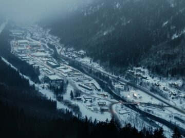 Aerial view of a snow-covered valley with a small town nestled among the mountains. Buildings, roads and trees are visible in the misty atmosphere and snow-covered landscape, where a Hyundai IONIQ 5 N makes its way through the serene winter scene.