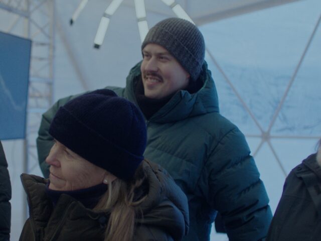 Three people, bundled in winter coats and hats, stand in a geometric tent structure, next to a sleek Hyundai IONIQ 5 N. They appear engaged and smiling, enjoying the snowy surroundings outside.