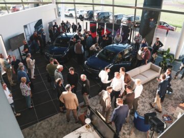 A busy Lancia Nederland showroom with groups of people examining new cars and interacting with salespeople.