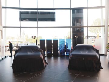 Twee met zwarte doeken beklede auto's in een moderne showroom met grote ramen, reflecterende vloer en een persoon op de achtergrond bij de eerste dealer.