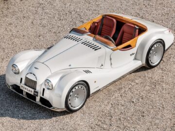 A white Morgan Midsummer Plus 8 GTR roadster with red leather seats and wood accents is parked on a gravel surface as seen from a high angle. This British-Italian beauty reflects timeless elegance and classic automotive craftsmanship.