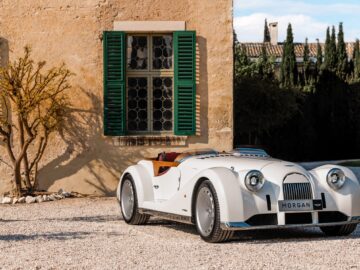 A vintage white Morgan sports car, emblematic of British-Italian beauty, is parked on a gravel driveway in front of a building with green shutters and a tree.