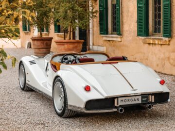 A white Morgan Midsummer convertible is parked on a gravel driveway in front of a yellow building with green shutters that epitomizes British-Italian beauty.