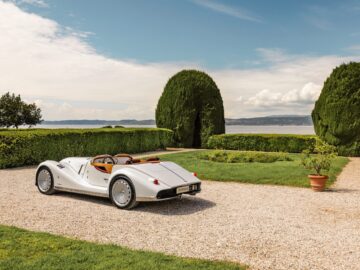 A white vintage-style Morgan Midsummer convertible is parked in a gravel driveway next to a neatly kept garden with hedgerows, overlooking a lake under a partly cloudy sky.
