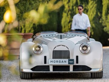 Een witte Morgan-sportwagen, die de Brits-Italiaanse schoonheid belichaamt, staat geparkeerd op een grindoprit met op de achtergrond een man met een zonnebril en een wit overhemd. Groen gebladerte is overal zichtbaar.