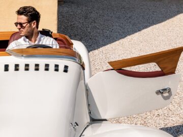 Een persoon met een zonnebril zit in een witte cabriolet met het bestuurdersportier open en straalt Brits-Italiaanse schoonheid uit.