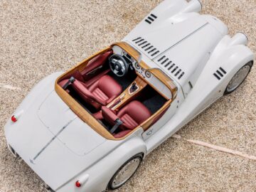 Aerial view of a vintage white convertible, a true British-Italian beauty with red leather seats and wooden dashboard, parked on a gravel surface.