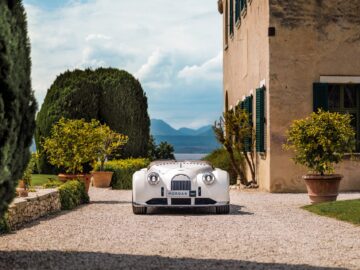 Een vintage zilveren Morgan Midsummer-sportwagen staat geparkeerd op een grindoprit naast een stenen gebouw met groene luiken, omgeven door verzorgde struiken en potplanten. De Brits-Italiaanse schoonheid zit tegen een achtergrond van bergen en blauwe lucht.