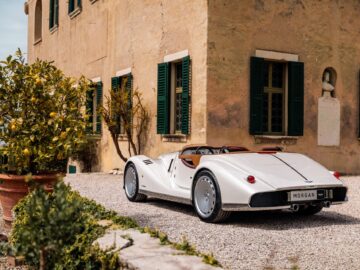 Die britisch-italienische Schönheit, ein weißer Morgan Midsummer Roadster im Vintage-Stil, parkt auf einer Kiesauffahrt neben einem beige verputzten Gebäude mit grünen Fensterläden und Topfpflanzen.
