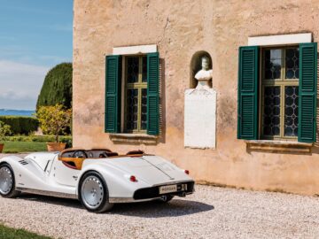 A British-Italian beauty, the Morgan Midsummer, a vintage-style white convertible, is parked in front of an old stone building with two windows with green shutters and a statue embedded in the wall.