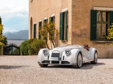 A beautiful white Morgan sports car, embodying British-Italian beauty, is parked on a gravel driveway next to a beige building with charming green shutters.